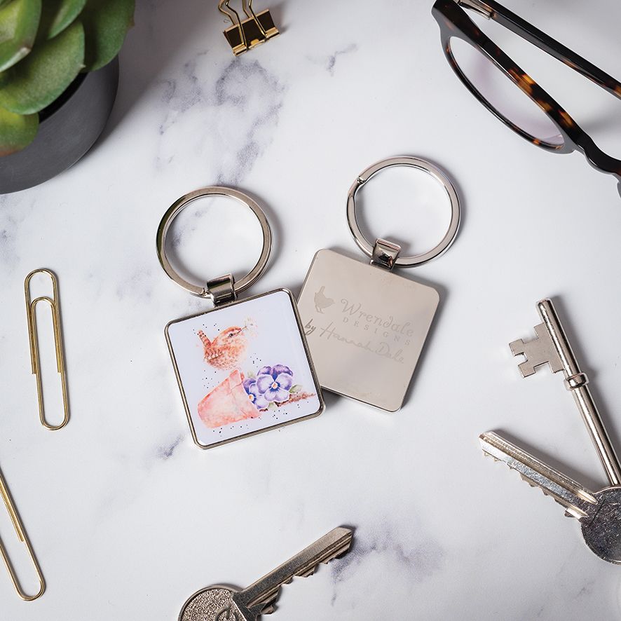 Two square metal keychains on a white marble surface with paper clips, keys, eyeglasses, and a succulent around them. One keychain has a printed floral design, and the other is engraved with the words 'Wishing you a Beautiful Day'.