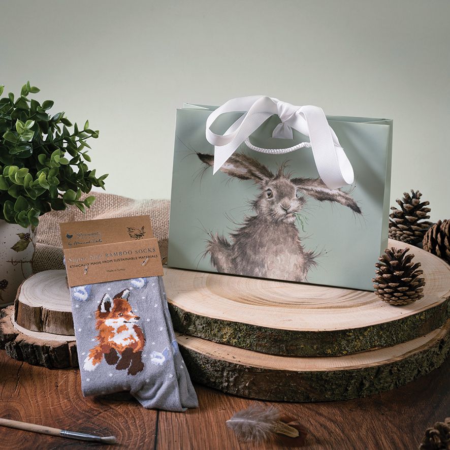 Gift bag with rabbit design and sock with fox design on a wooden surface with plants and pinecones.