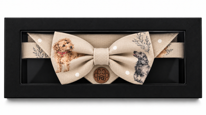 A beige fabric bow tie displayed inside a black rectangular presentation box. The bow tie features printed dog illustrations, white polka dots, and a central round wooden medallion with a paw print and the initials 'FB'.