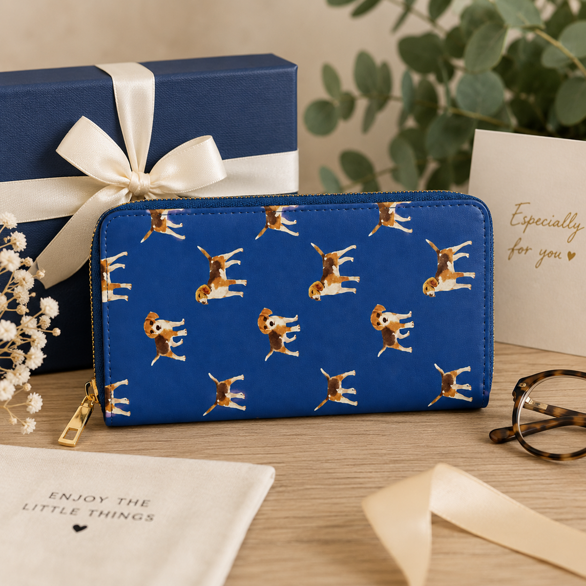 A blue zip-around wallet with a repeating pattern of Beagle dogs, styled on a wooden table next to a blue gift box with a white ribbon, a card that says 'Especially for you', and a pair of eyeglasses.