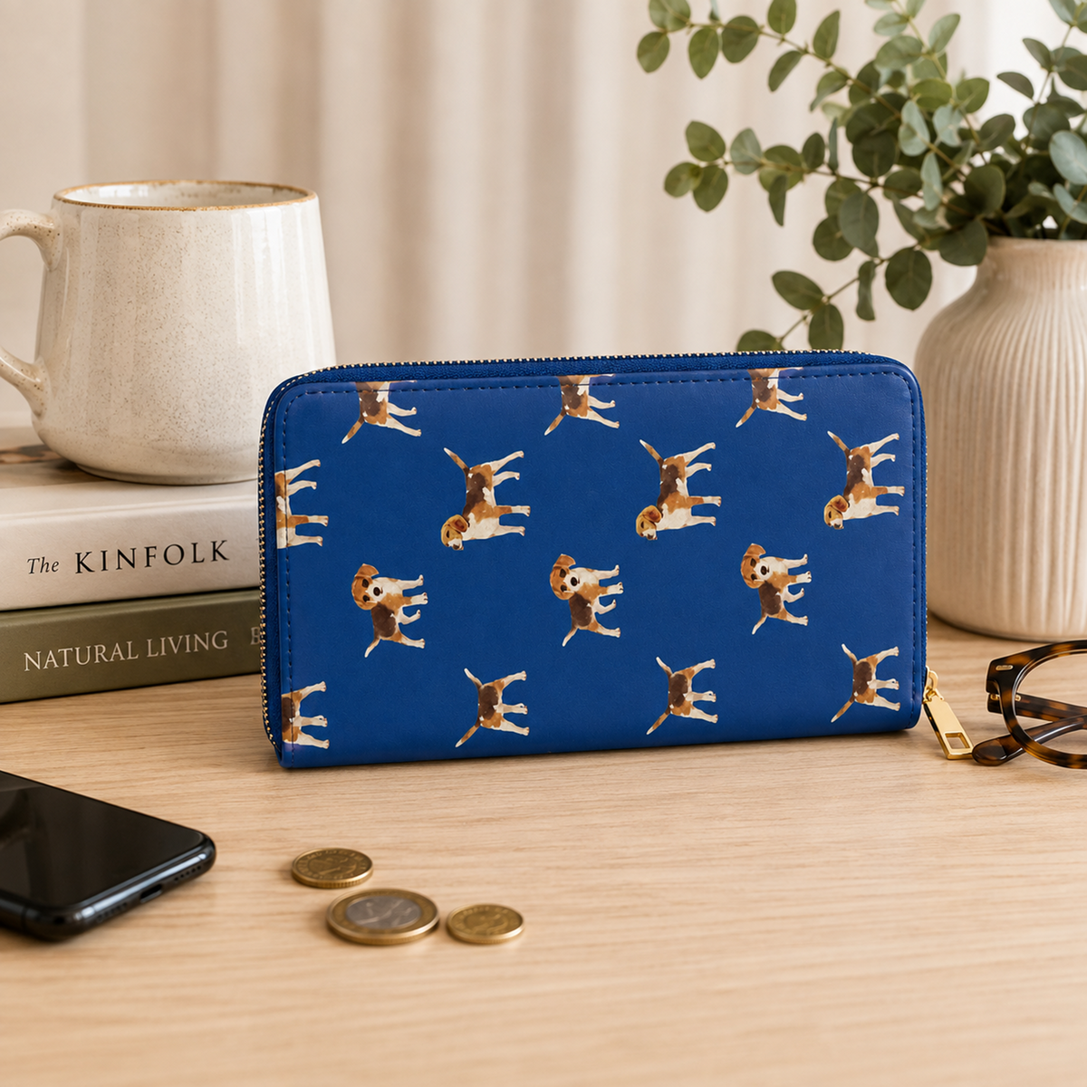 A blue zip-around wallet with a repeating print of Beagle dogs lying on their backs, styled on a wooden tabletop with a mug, books, coins, a phone, and eyeglasses nearby.