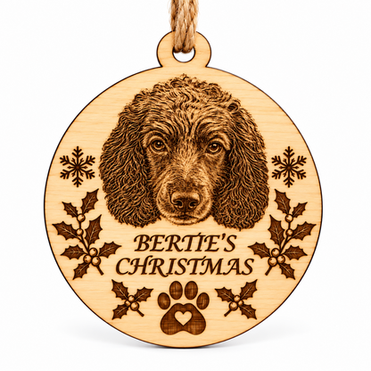 A round wooden hanging ornament engraved with a dog portrait, holly leaves, snowflakes, a paw print, and the text 'BERTIE'S CHRISTMAS,' suspended by a natural twine loop against a clean white background.