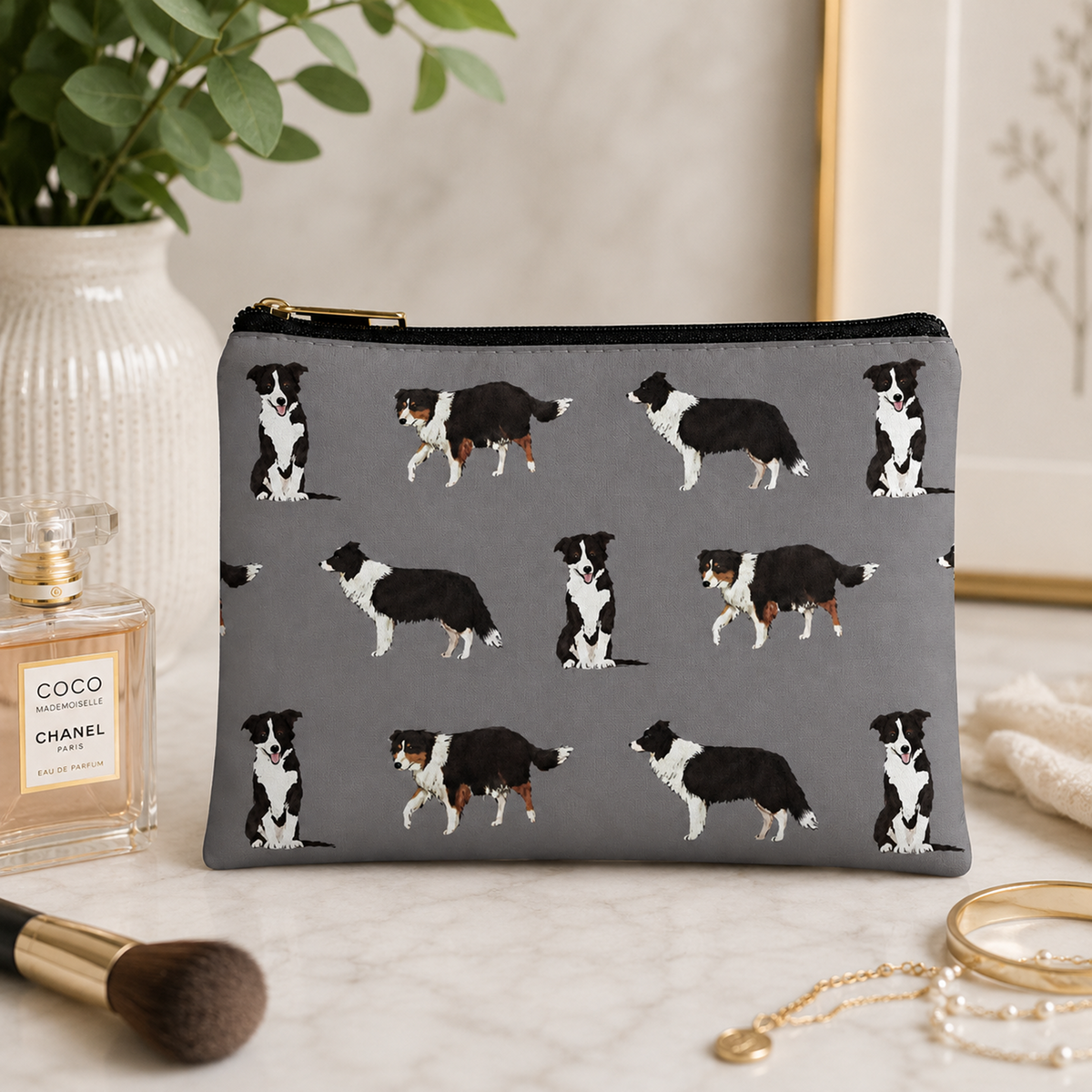 A lifestyle image of a grey zipped cosmetic bag featuring a repeating pattern of black and white Border Collie dogs, with a gold zipper pull.