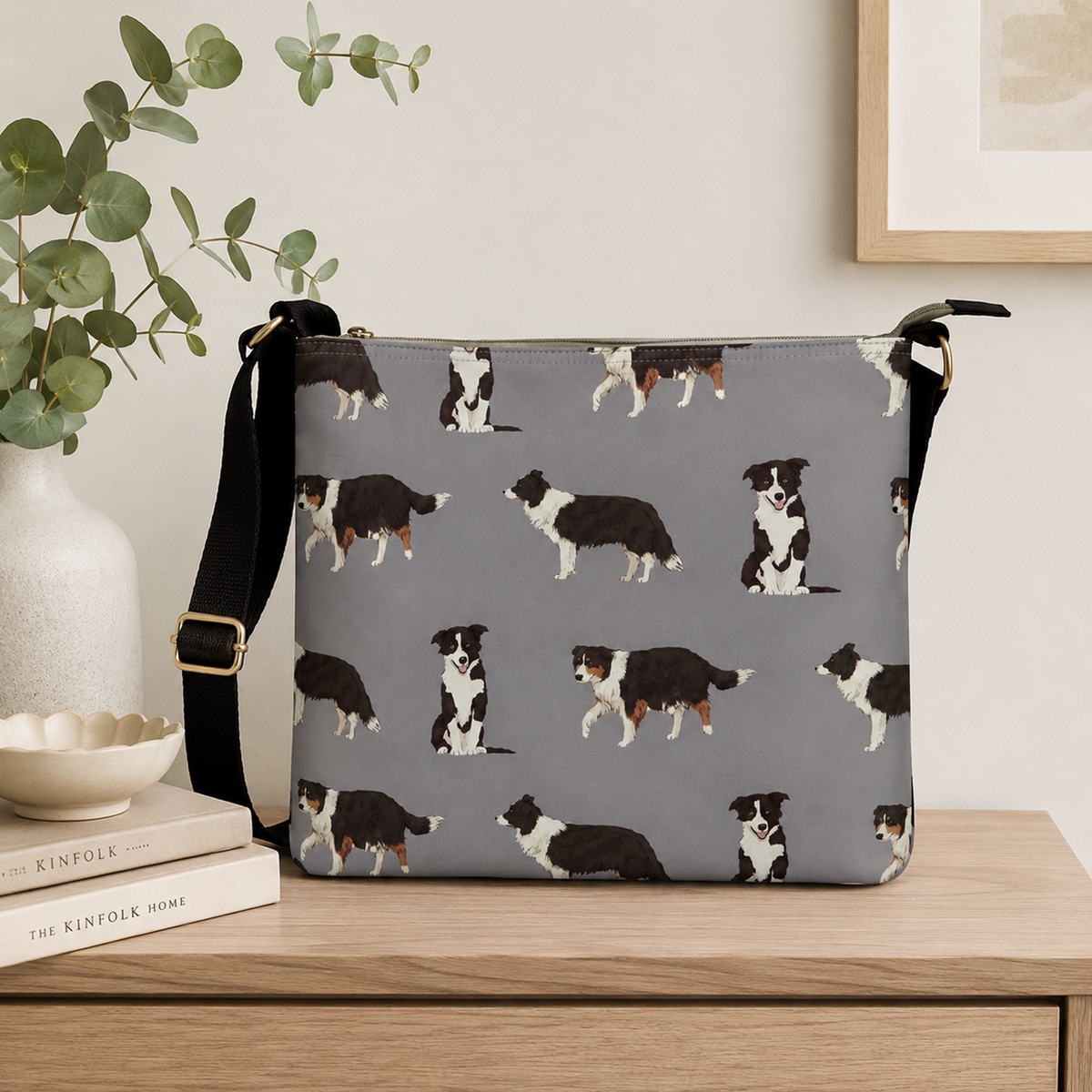 A grey shoulder bag with a repeating pattern of black, white, and brown Border Collie dogs, styled on a wooden table next to a vase with eucalyptus and books.