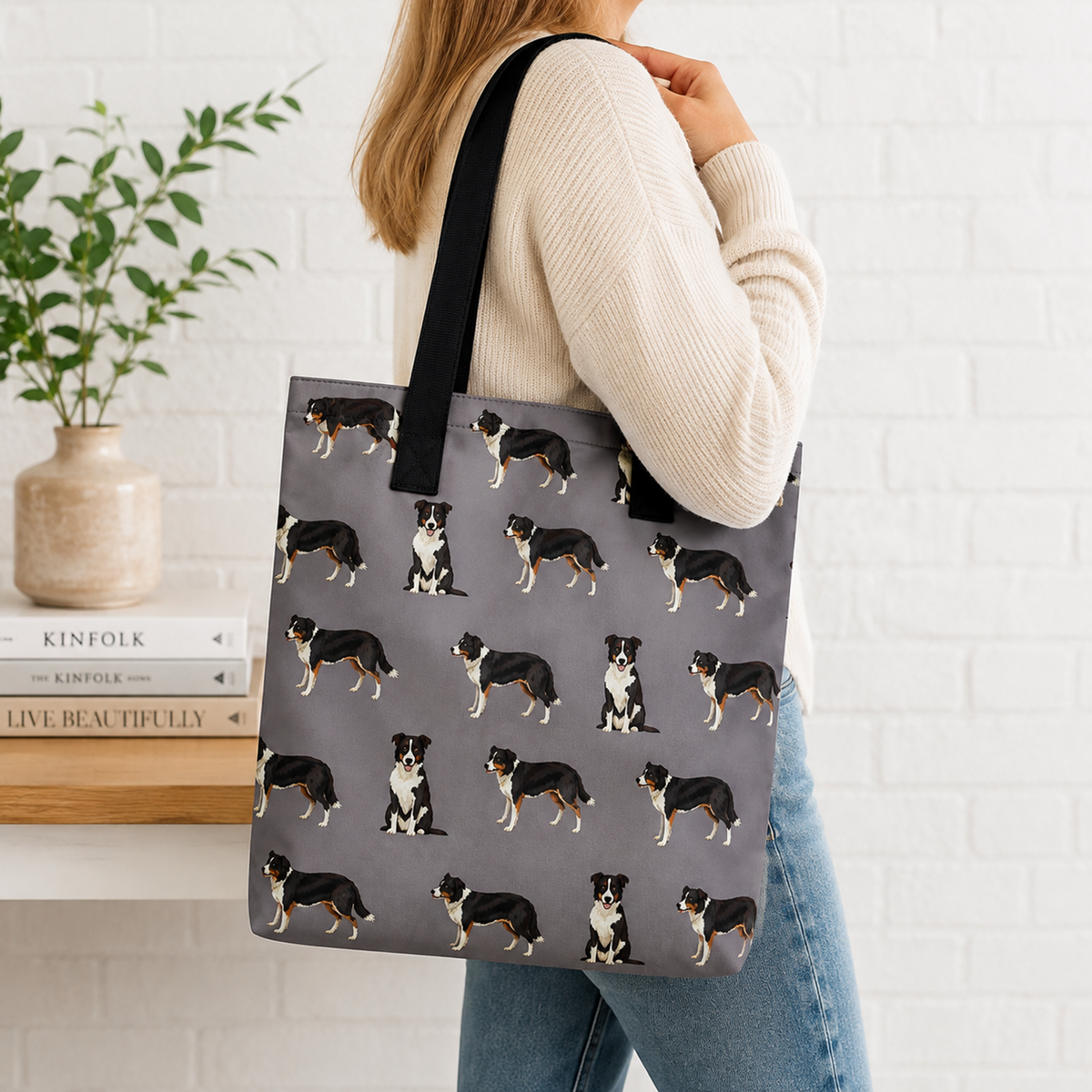 A woman wearing a cream ribbed sweater and blue jeans carries a grey tote bag with a black shoulder strap. The tote bag features an all-over print of black, white, and tan dogs.