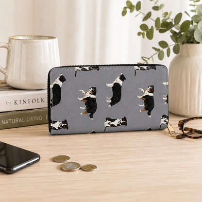 A grey zip-around wallet with a repeating print of black-and-white Border Collie dog figures, styled on a light wood tabletop with a mug, book, phone, coins, glasses, and a vase in the background.