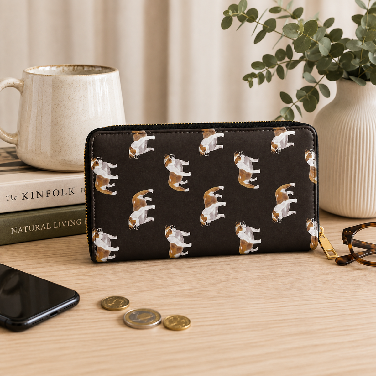 A black zip-around wallet with a repeating print of white, brown, and tan British Bulldogs, styled on a light wood desk with a mug, books, phone, coins, and eyeglasses nearby.