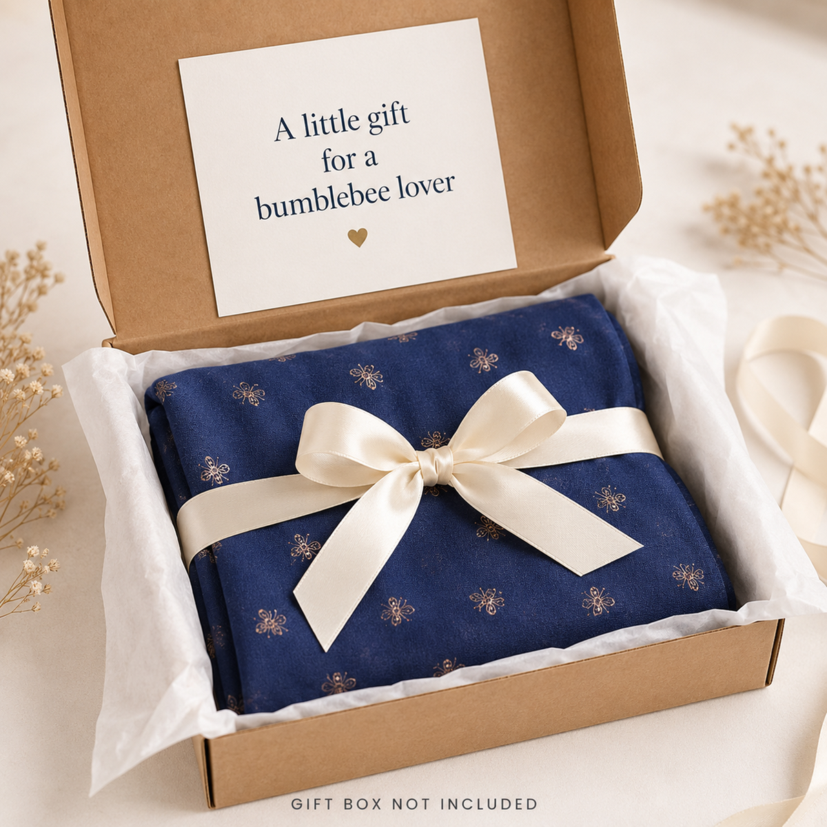 A denim blue gift-wrapped package tied with a cream ribbon, presented inside an open kraft gift box with a white card that reads 'A little gift for a bumblebee lover.' The box is styled on a light surface with dried floral props around it.