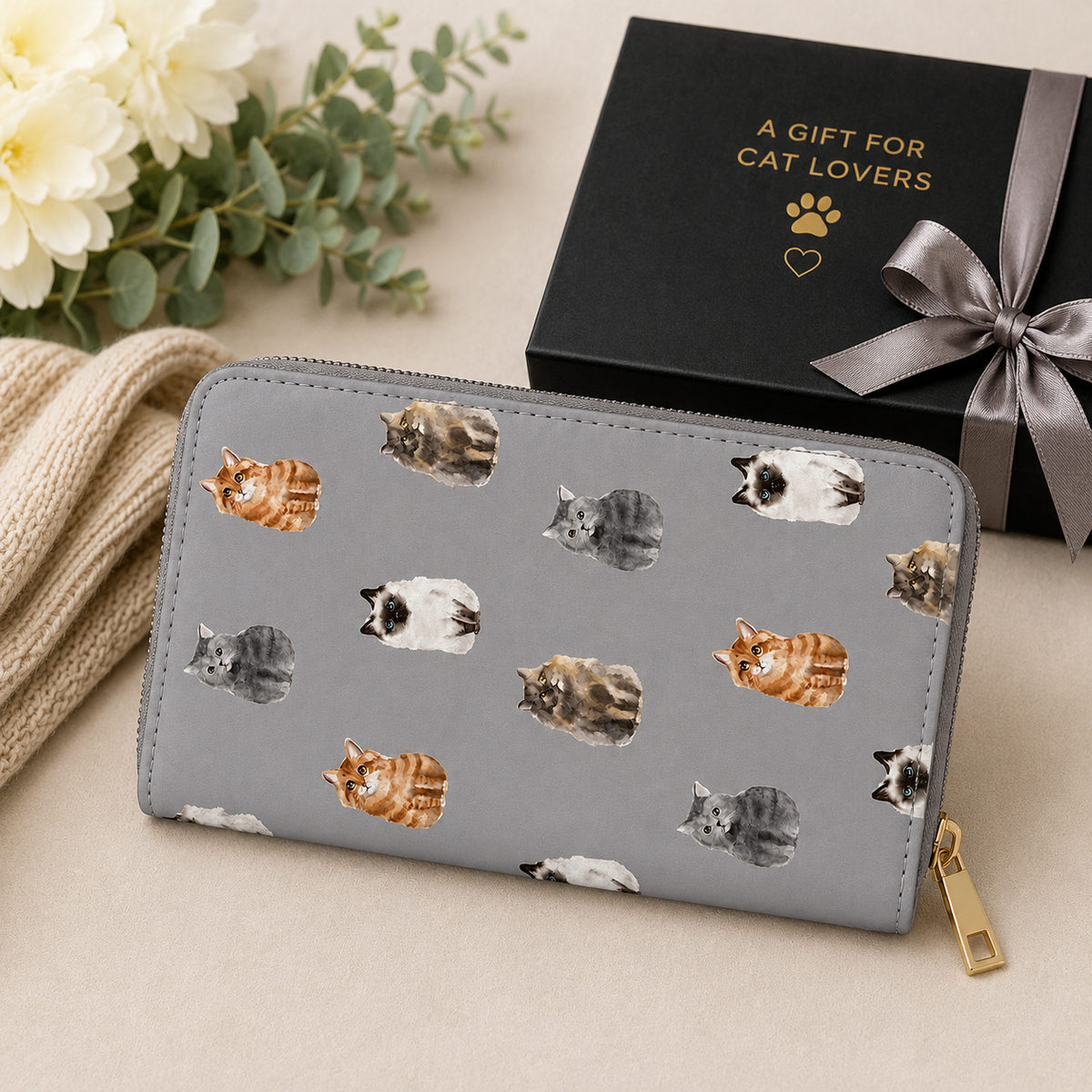 A grey zip-around wallet with a print of various cat illustrations, styled on a beige surface next to a black gift box with gold lettering and a grey ribbon.