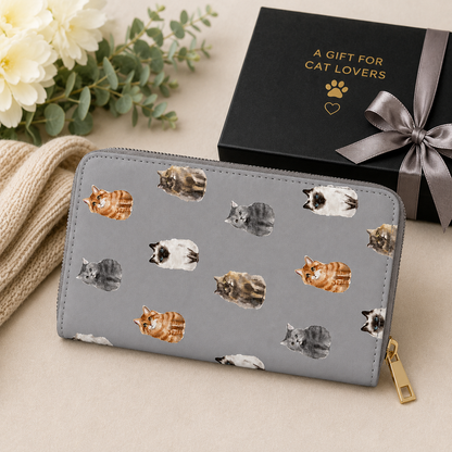 A grey zip-around wallet with a print of various cat illustrations, styled on a beige surface next to a black gift box with gold lettering and a grey ribbon.
