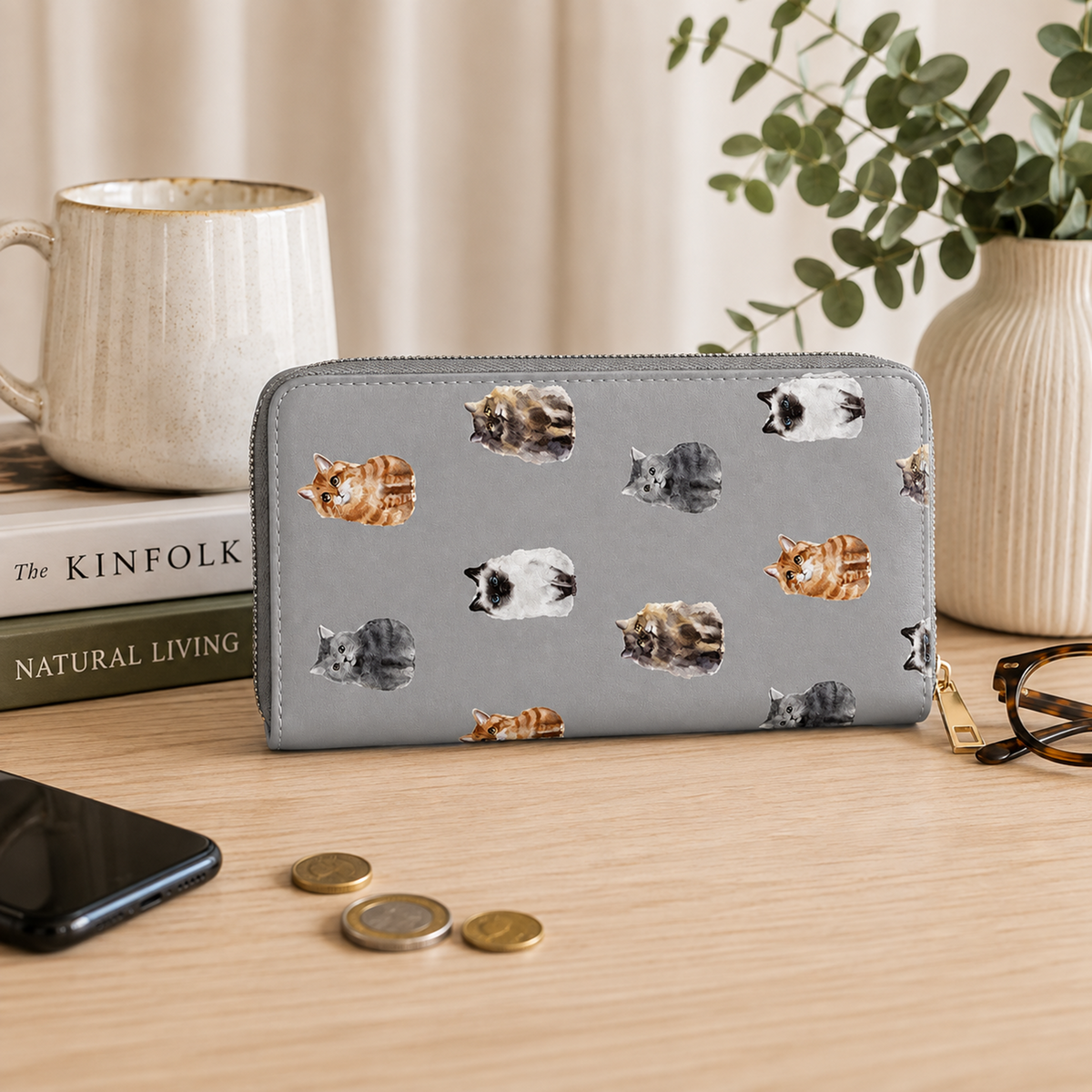 A grey zip-around wallet with a repeating pattern of illustrated cats in orange, black, white, and grey, displayed on a wooden tabletop with a mug, books, phone, coins, and eyeglasses nearby.