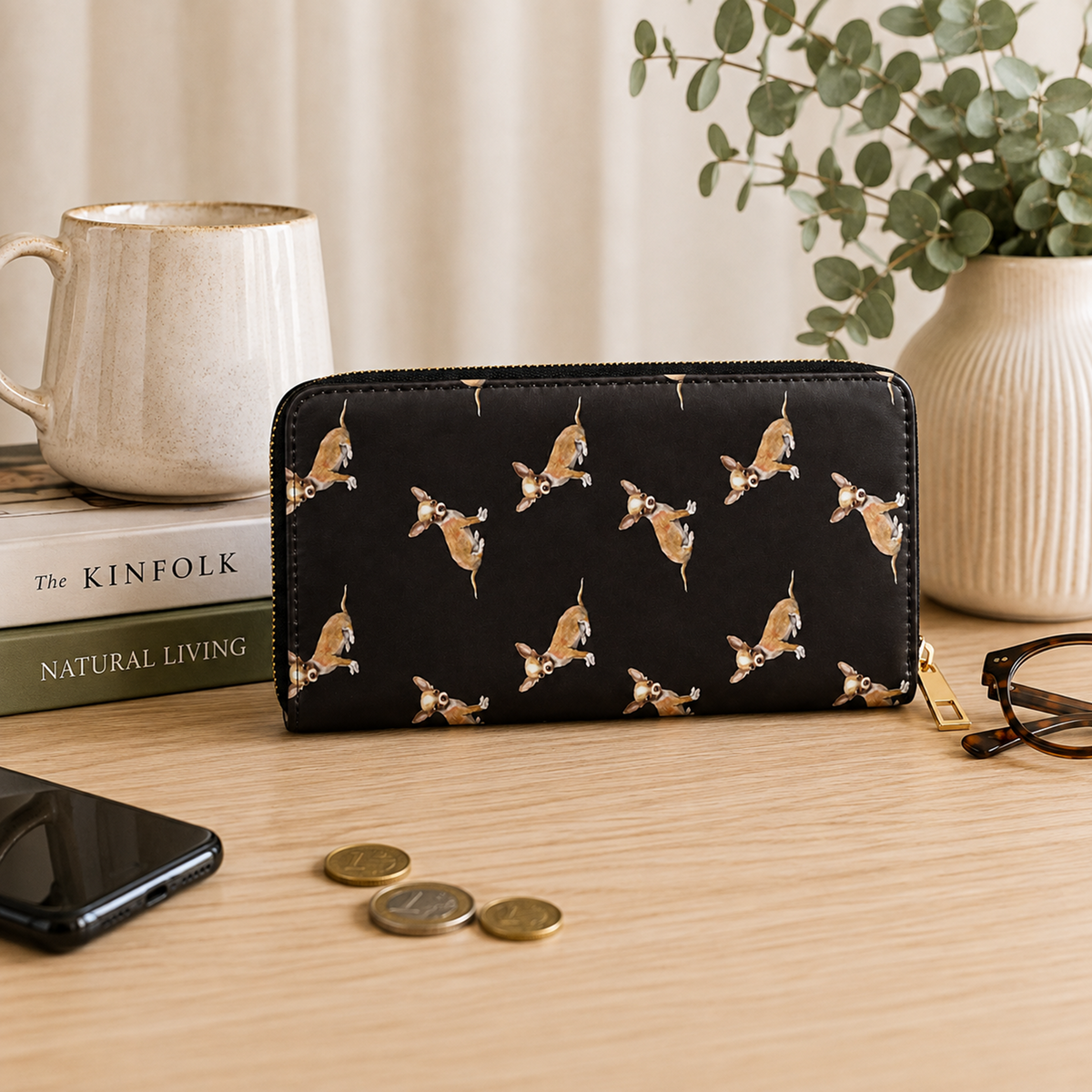 A black zip-around wallet with a repeating print of small tan and white Chihuahua dogs, styled on a light wood tabletop with a mug, books, coins, a phone, eyeglasses, and a vase of greenery in the background.