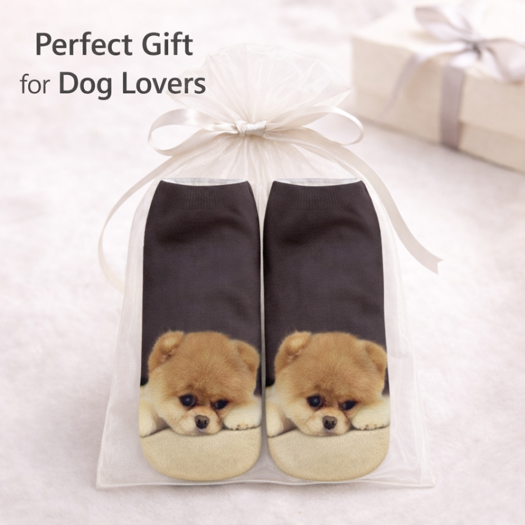 A pair of dark charcoal socks with printed fluffy cream and tan Chow Chow dog faces on the soles, presented in a clear tulle gift bag tied with a white ribbon. Text in the upper left reads 'Perfect Gift for Dog Lovers' on a soft white background with a blurred gift box in the background.