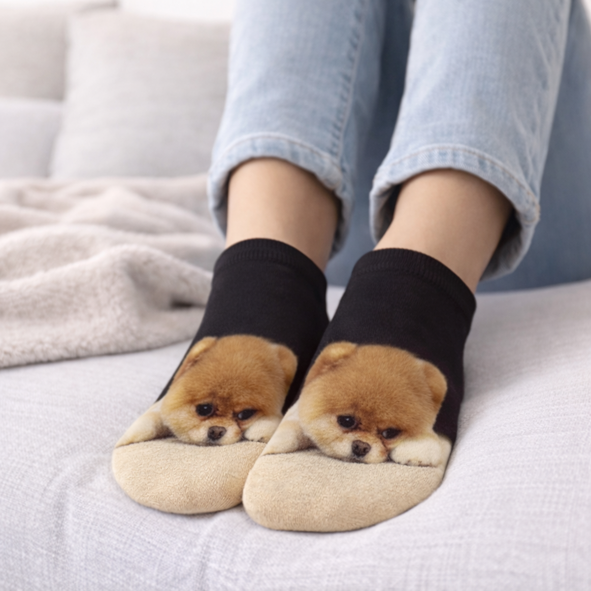 A person wearing black trainer socks with a printed fluffy brown Chow Chow dog face on the toes, standing on a light-coloured bed or sofa.