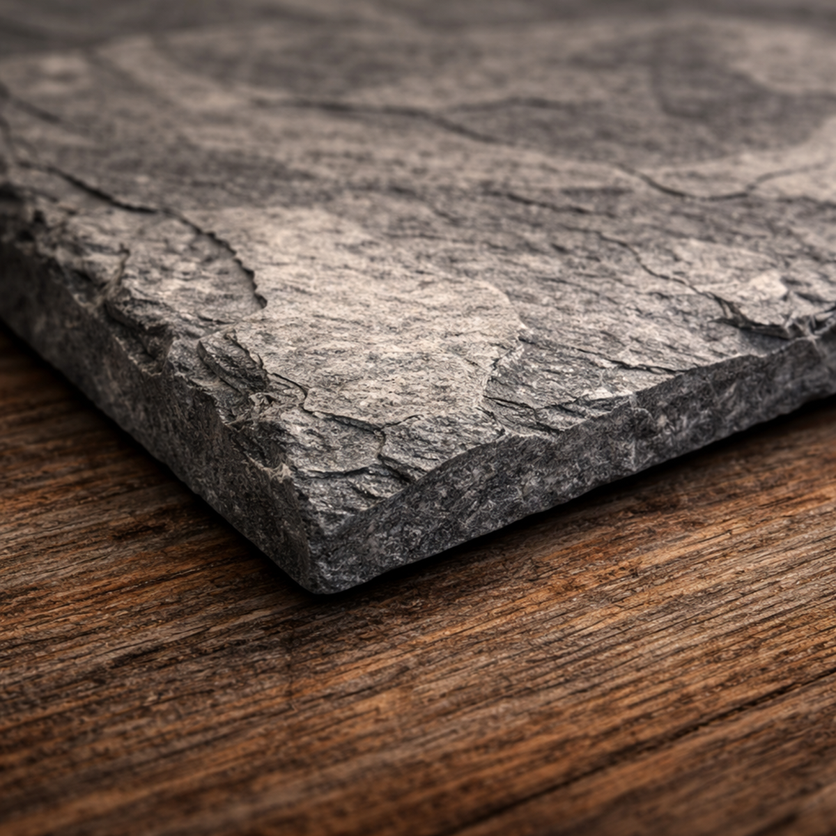 A close-up of a dark gray slate stone tile with a rough, textured surface, resting on a rustic brown wooden surface.