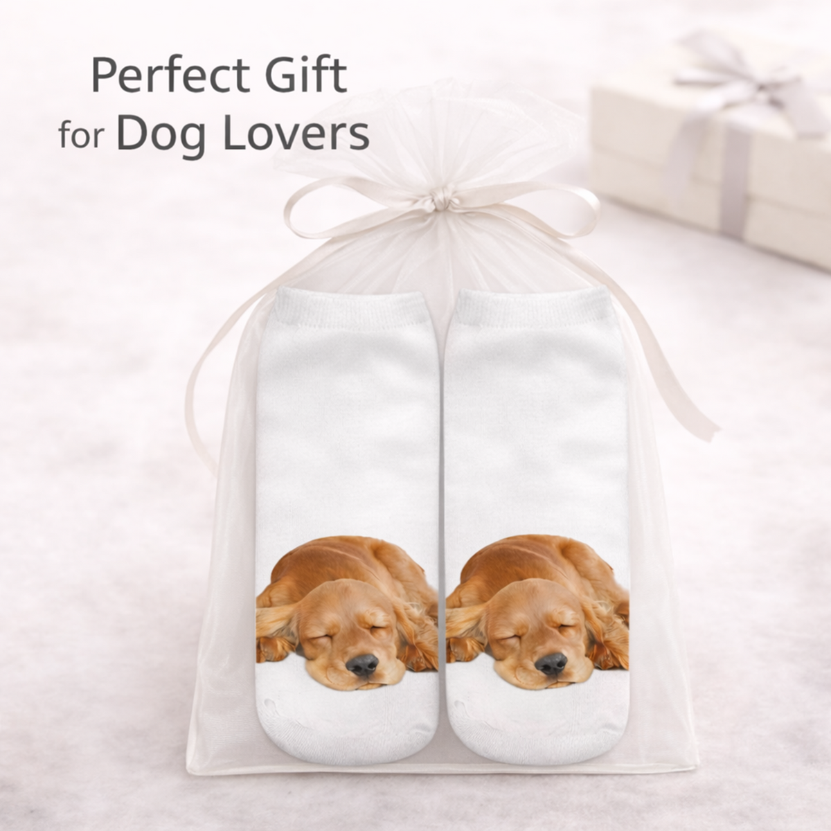A gift set containing a pair of white socks printed with a sleeping Cocker Spaniel dog, presented in a clear mesh gift bag tied with a white ribbon. The text 'Perfect Gift for Dog Lovers' is displayed at the top, with a blurred gift box in the background.