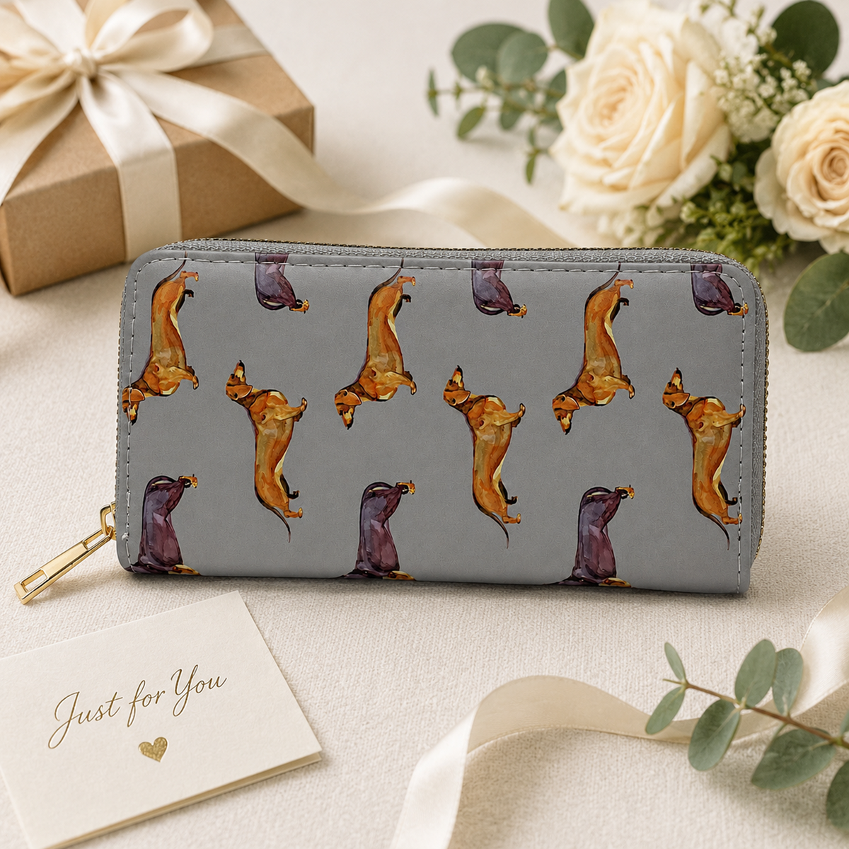 A gray zip-around wallet with a repeating pattern of dachshund dogs in orange, brown, and purple, styled on a beige fabric surface with a gift box, cream roses, eucalyptus, and a small card reading 'Just for You'.