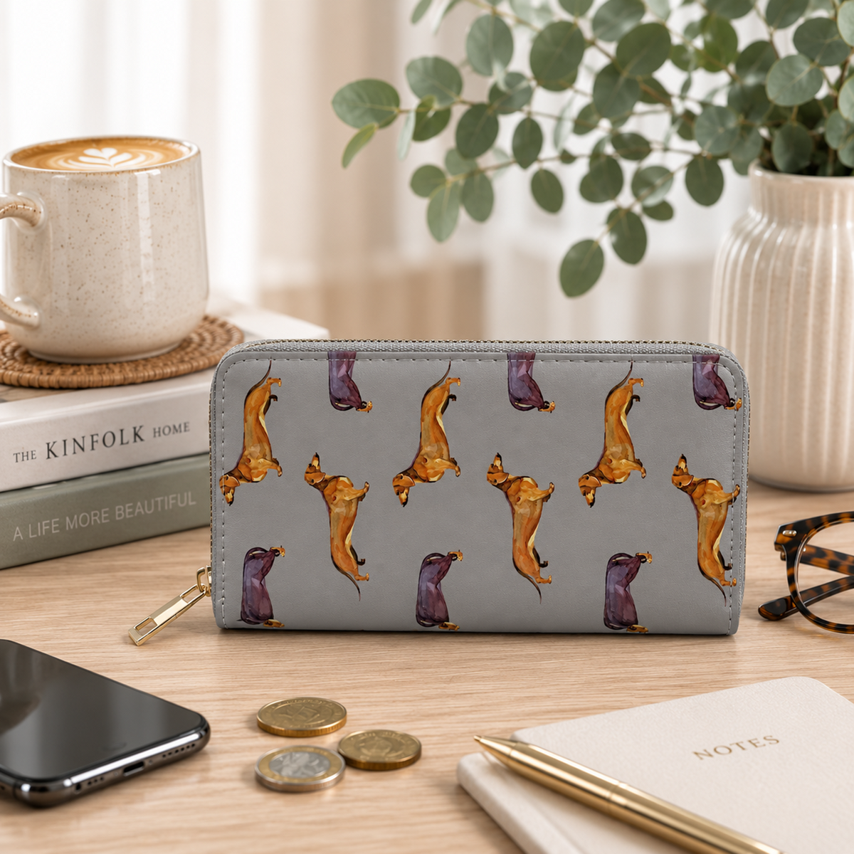 A gray zip-around wallet with a repeating dachshund print, styled on a wooden desk with a coffee cup, books, phone, coins, pen, and glasses.