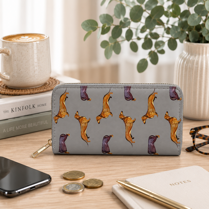 A gray zip-around wallet with a repeating dachshund print, styled on a wooden desk with a coffee cup, books, phone, coins, pen, and glasses.