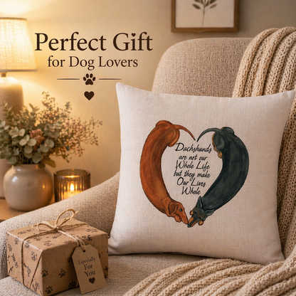 A square cushion cover pillow in a cosy living room setting, featuring a printed design of two dachshunds forming a heart shape with the text 'Dachshunds are not our Whole Life but they make Our Lives Whole.' A wrapped gift box and a lamp are visible in the background, with the text 'Perfect Gift for Dog Lovers' overlaid on the image.