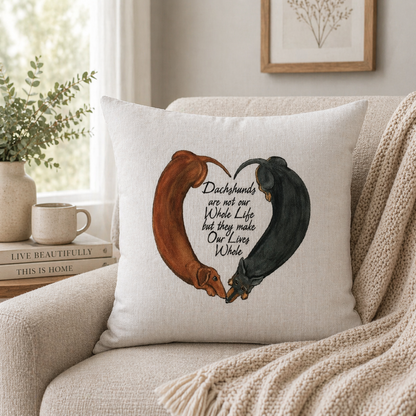 A square cushion cover pillow in a cosy living room setting, featuring an illustration of two dachshunds forming a heart shape with the text 'Dachshunds are not our Whole Life but they make Our Lives Whole'.