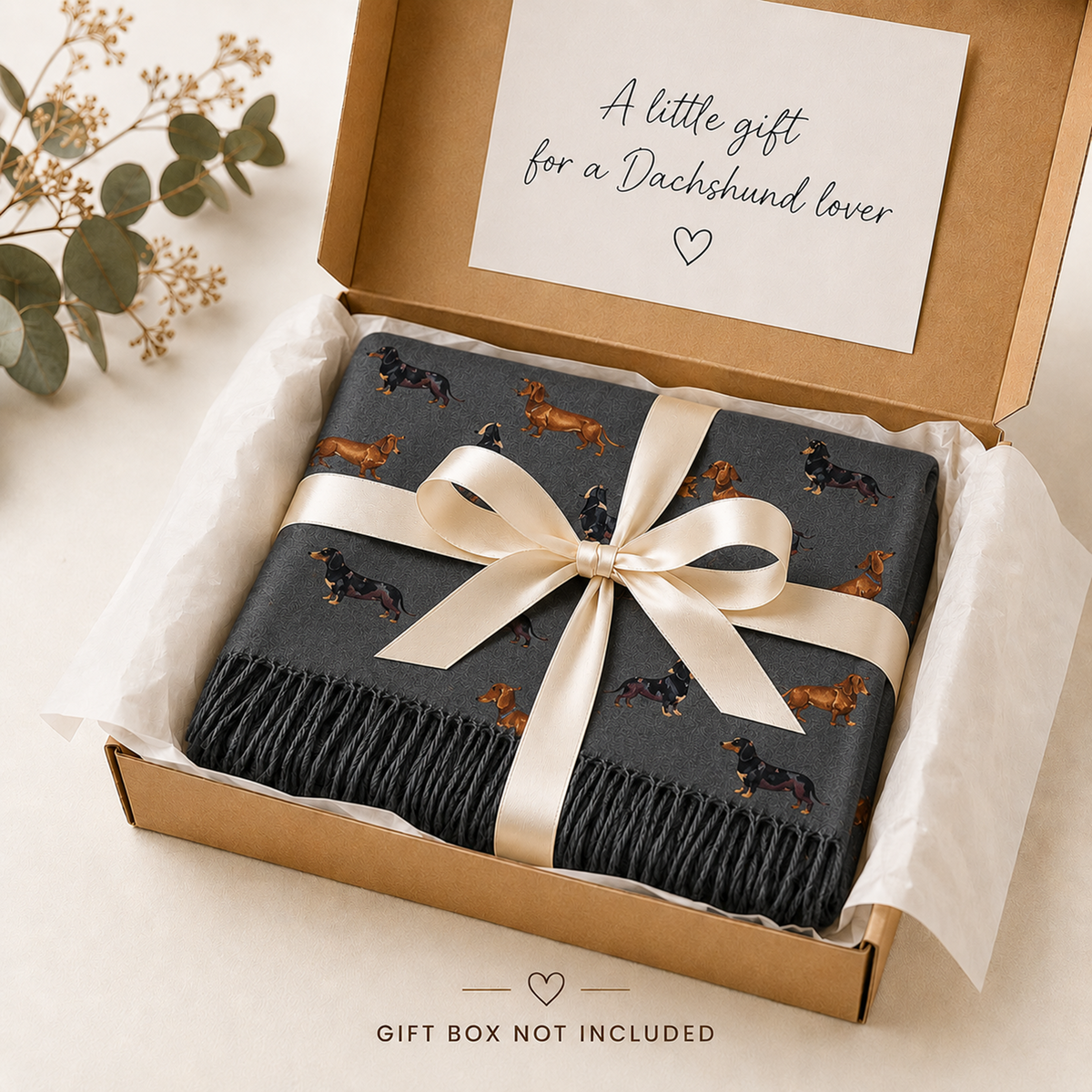 A folded dark charcoal grey scarf with a dachshund print, tied with a cream satin ribbon and presented in a brown gift box with a handwritten note inside. A small eucalyptus sprig decorates the top left corner.