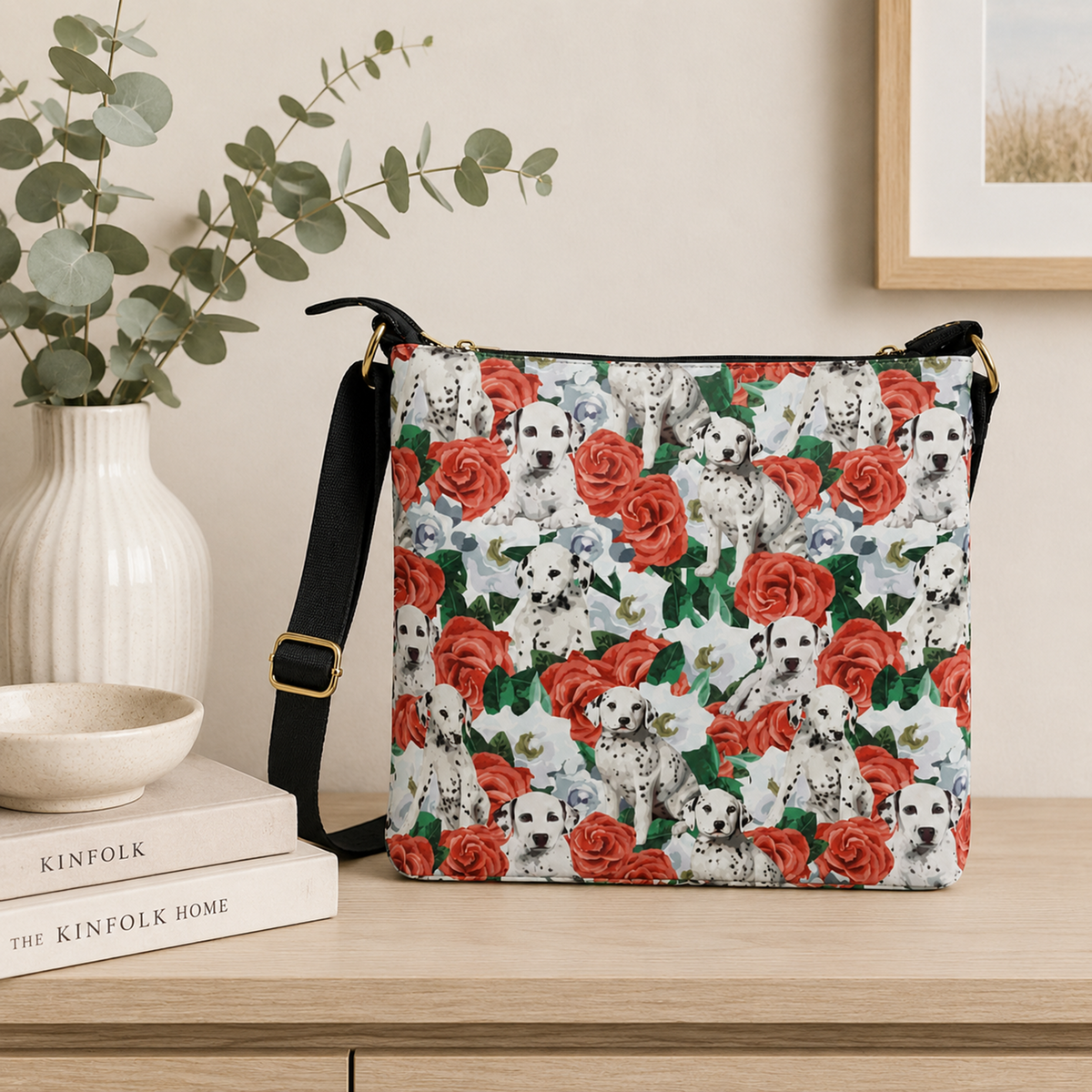 A crossbody handbag with a repeating pattern of Dalmatian puppies and red roses, resting on a light wood table next to a white ceramic vase with eucalyptus branches.