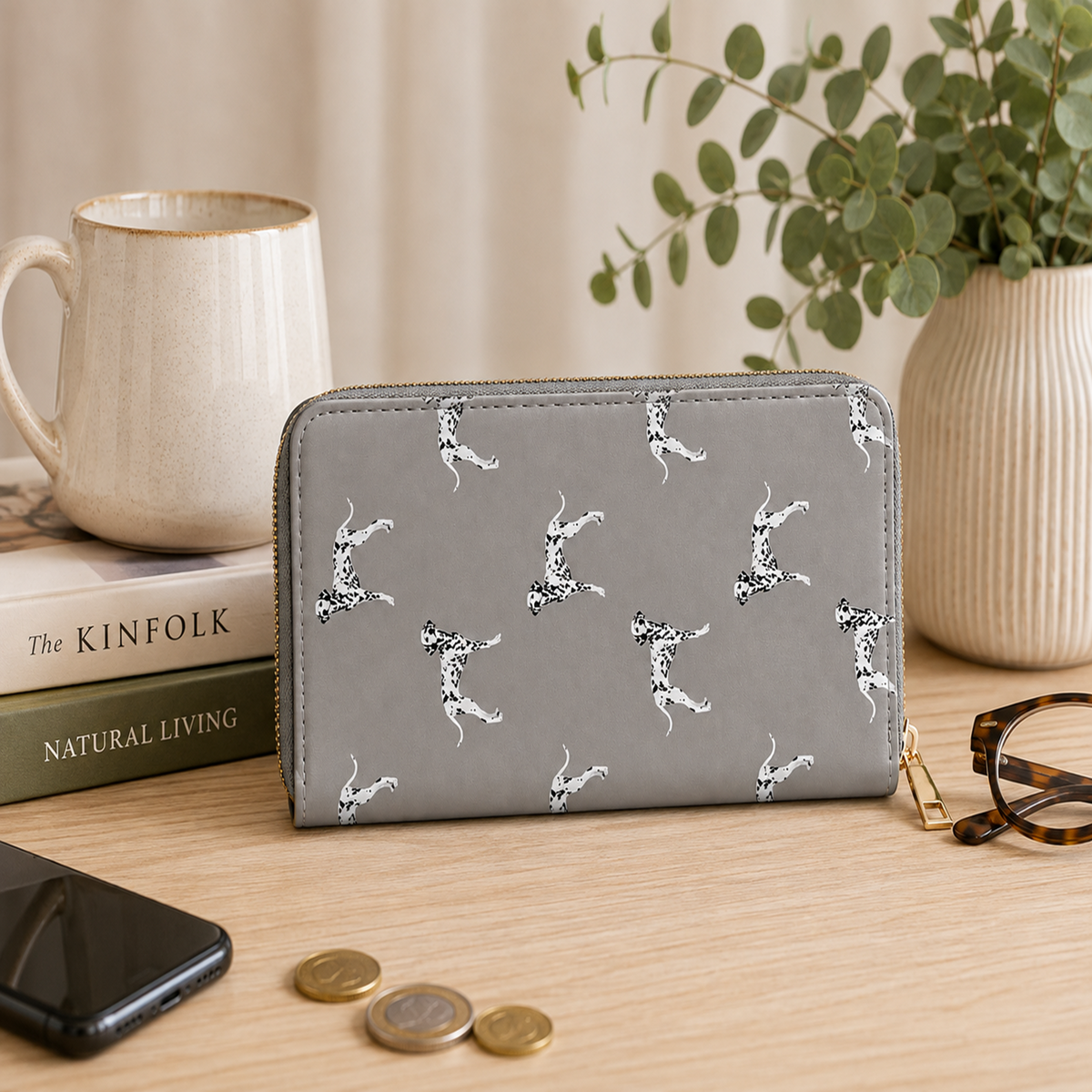 A grey zip-around wallet with a white and black dalmatian dog print pattern, styled on a wooden desk with a mug, books, a phone, coins, eyeglasses, and a potted plant in the background.