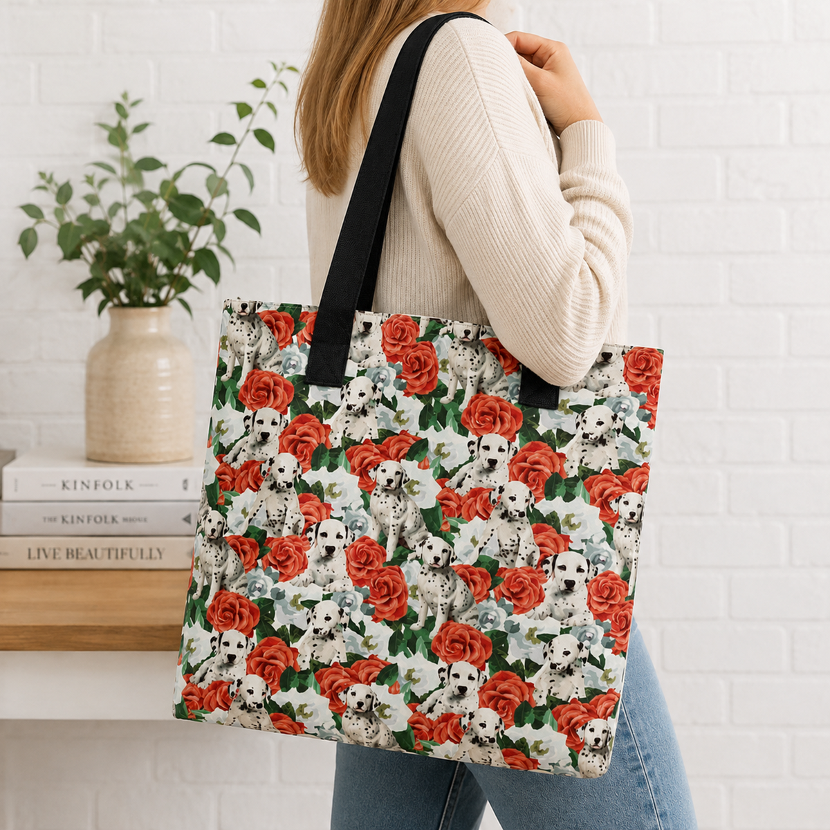 A woman wearing a cream ribbed sweater and blue jeans carries a large tote bag with a repeating pattern of white Dalmatian dogs and red roses with green leaves. The background shows a white brick wall, a potted plant, and books.
