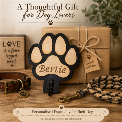 Personalised wooden dog lead holder plaque shaped like a paw print, shown with a brown gift box, rope leash, collar, and decorative text on a rustic tabletop.