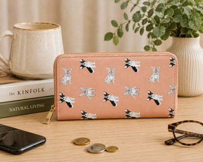 A pink zip-around wallet with a repeating black-and-white French Bulldog dog print, styled on a wooden tabletop with a mug, books, plant, phone, coins, and glasses around it.