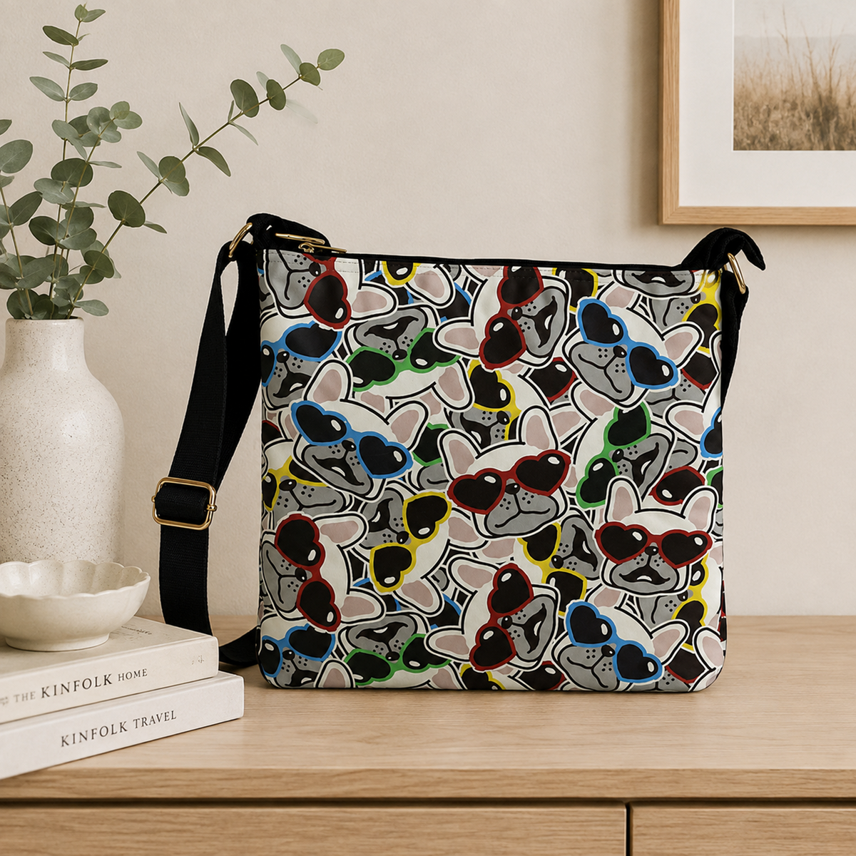 A rectangular shoulder bag with a colorful cartoon dog print wearing sunglasses, styled on a wooden table next to a white vase with eucalyptus and a stack of books.