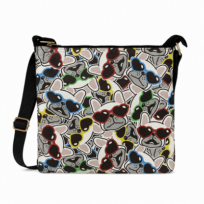 A crossbody bag with a white background and an all-over pattern of cartoon French bulldogs wearing colorful sunglasses. The bag has a black shoulder strap with a gold buckle and gold zipper hardware.