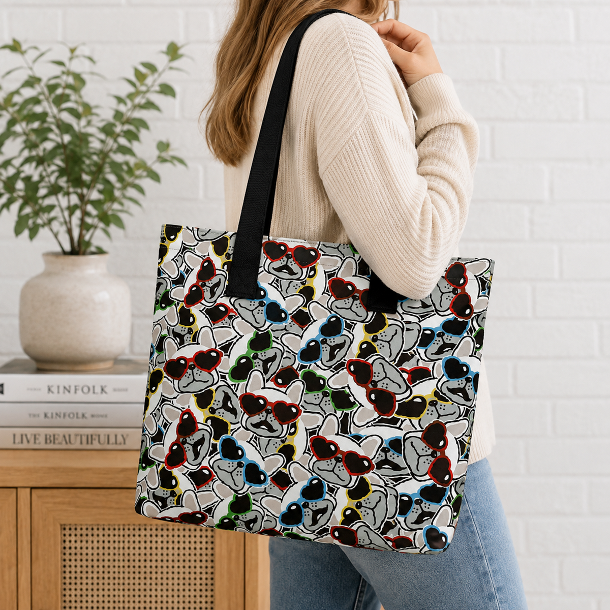 A woman wearing a cream ribbed sweater and blue jeans carries a large tote bag with a black handle. The tote bag features an all-over print of stylized cartoon French Bulldogs in multiple colours on a white background.