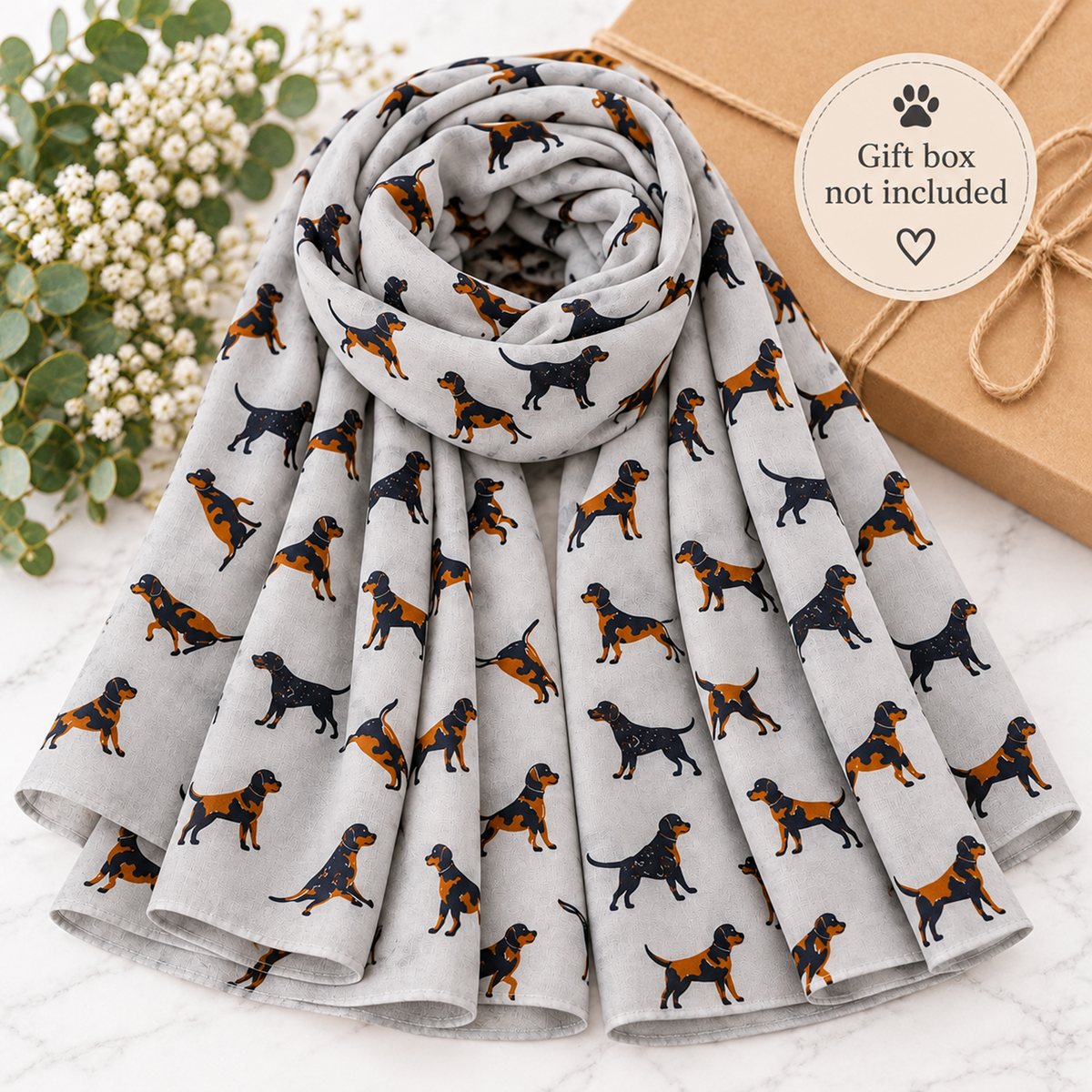 A light grey fabric gift box wrap or scarf featuring a repeating print of black and tan dogs, styled on a white marble surface next to white flowers and a kraft gift box with a 'Gift box not included' sticker.
