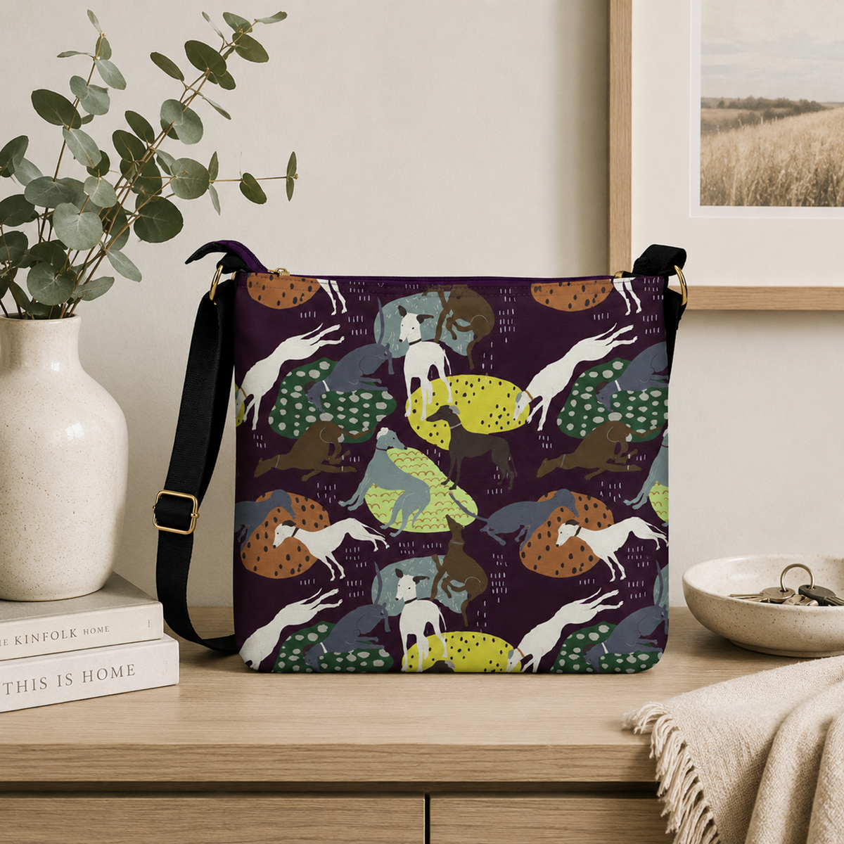 A rectangular shoulder bag with a dark purple background and an all-over whimsical Greyhound dog pattern in white, yellow, green, and brown, displayed on a wooden table in a styled home interior next to a vase of eucalyptus.