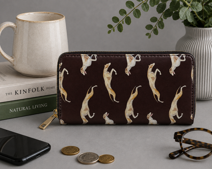 A dark brown zip-around wallet with a repeating pattern of leaping tan and white Greyhound & Whippet dogs, styled on a grey tabletop with a ceramic mug, books, a smartphone, coins, eyeglasses, and a potted plant.