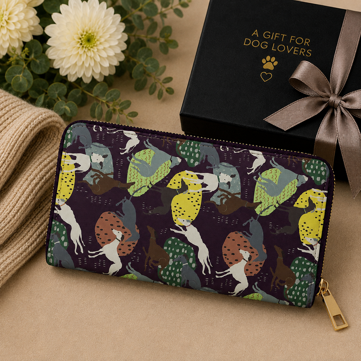 A long zip-around wallet with a colourful Greyhound and Whippet dog-and-woodland pattern, styled on a beige surface next to a black gift box with gold text and a ribbon, with flowers and a knit sweater in the background.