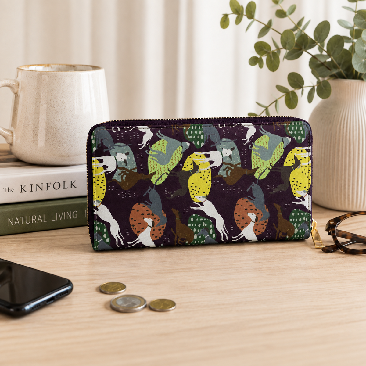 A zip-around wallet with a colourful Greyhound & Whippet dog pattern, styled on a wooden tabletop next to a smartphone, coins, eyeglasses, a mug, and books.