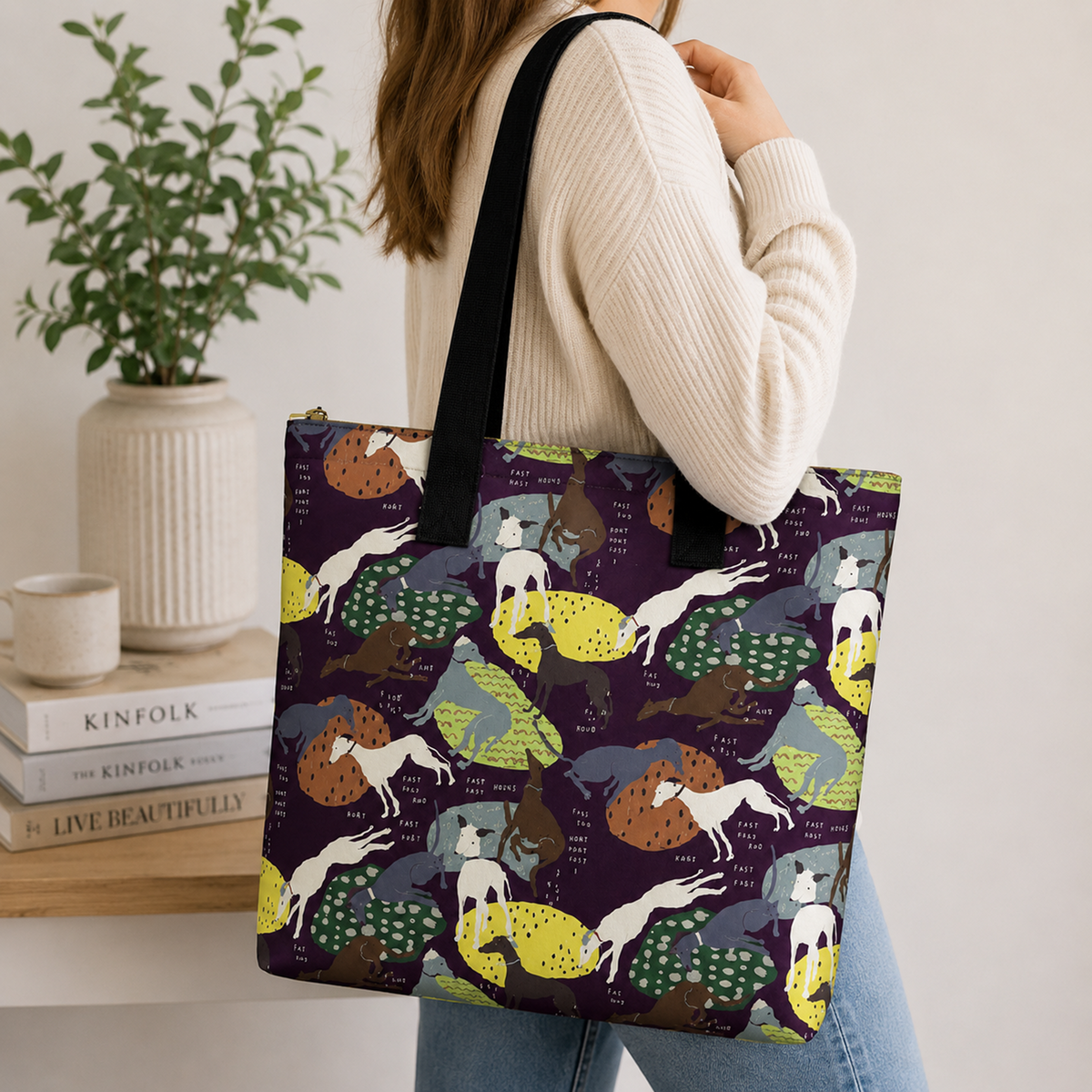 A woman wearing a cream ribbed sweater and blue jeans carries a large tote bag over her shoulder. The tote bag features a dark purple background with a whimsical pattern of Greyhound Whippet dogs in yellow, green, brown, and white, along with small text labels.