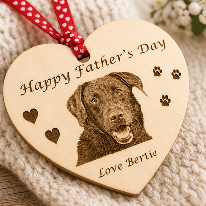 A heart-shaped wooden Father's Day ornament engraved with a dog portrait, paw prints, and the text 'Happy Father's Day' and 'Love Bertie,' hanging from a red ribbon with white hearts, styled on a cream knit blanket.