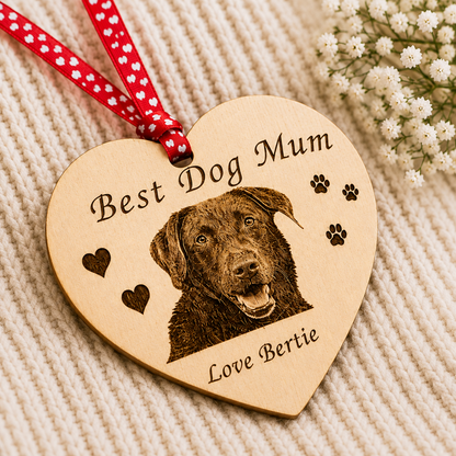 A heart-shaped wooden dog memorial ornament engraved with 'Best Dog Mum' and 'Love Bertie' alongside a portrait of a brown dog, hanging from a red and white polka-dot ribbon on a cream knit blanket with small white flowers in the corner.