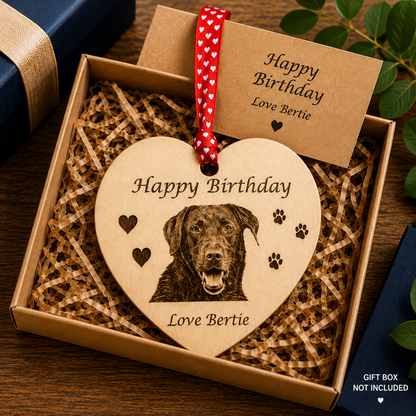 A heart-shaped wooden pet memorial ornament engraved with a dog portrait and the words 'Happy Birthday Love Bertie,' displayed in a gift box with shredded paper. A kraft card reading 'Happy Birthday Love Bertie' is tucked behind it, and part of a navy gift box is visible at the edges.