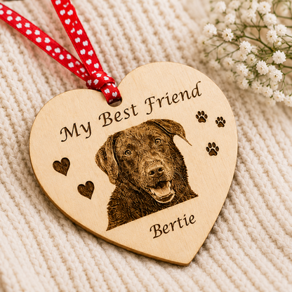 A heart-shaped wooden pet memorial ornament engraved with 'My Best Friend' and the name 'Bertie' alongside a photo of a chocolate Labrador retriever. The ornament hangs from a red ribbon with white polka dots and is displayed on a cream-colored knit blanket with small white flowers in the background.