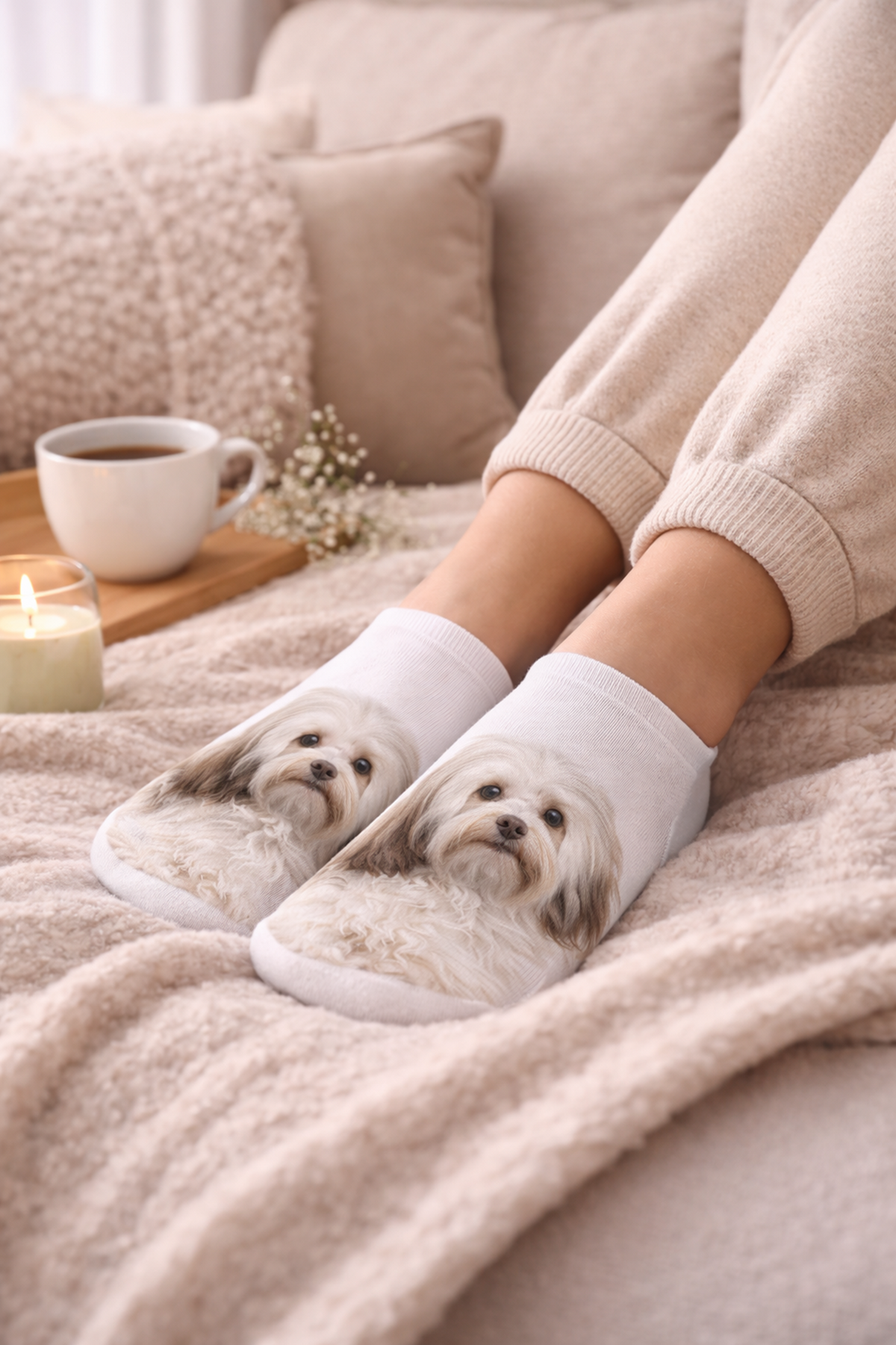 A person reclining on a beige blanket with pillows, wearing white trainer socks printed with a fluffy Havanese dog face. A cup of coffee, a candle, and small flowers sit nearby in a cosy indoor setting.