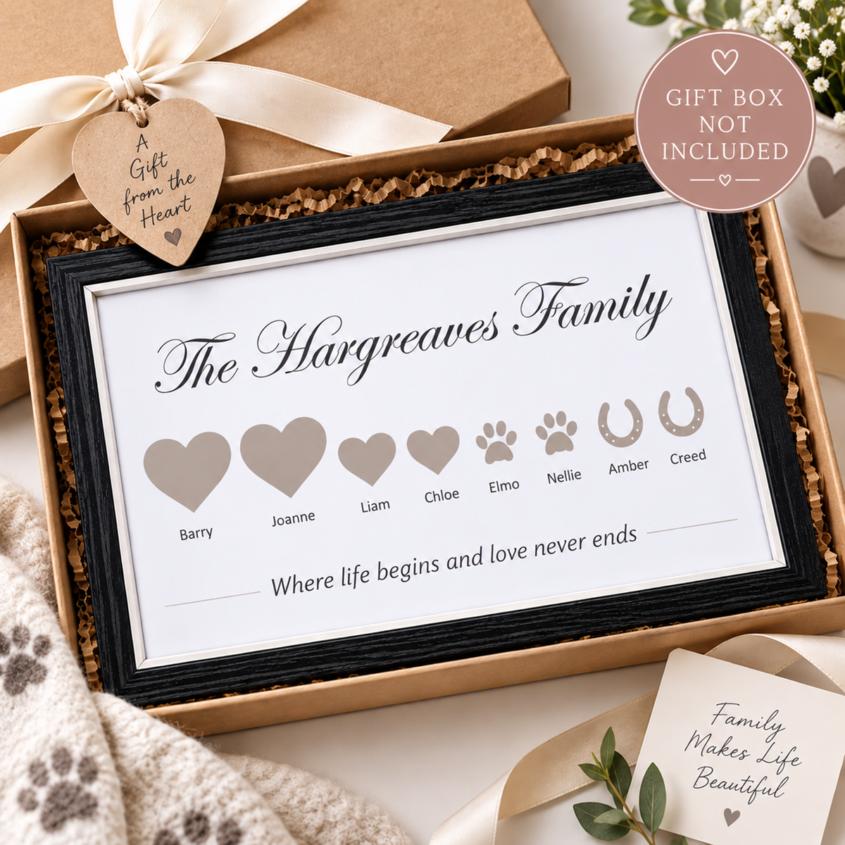 A personalised family memorial print in a black frame, presented inside a kraft gift box with a cream ribbon and heart tag. The print features customizable heart and paw print names under the heading 'The Hargreaves Family' with the quote 'Where life begins and love never ends'.