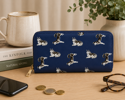 A navy blue zip-around wallet with a repeating print of husky dogs, styled on a wooden desk with a mug, books, phone, coins, and eyeglasses.