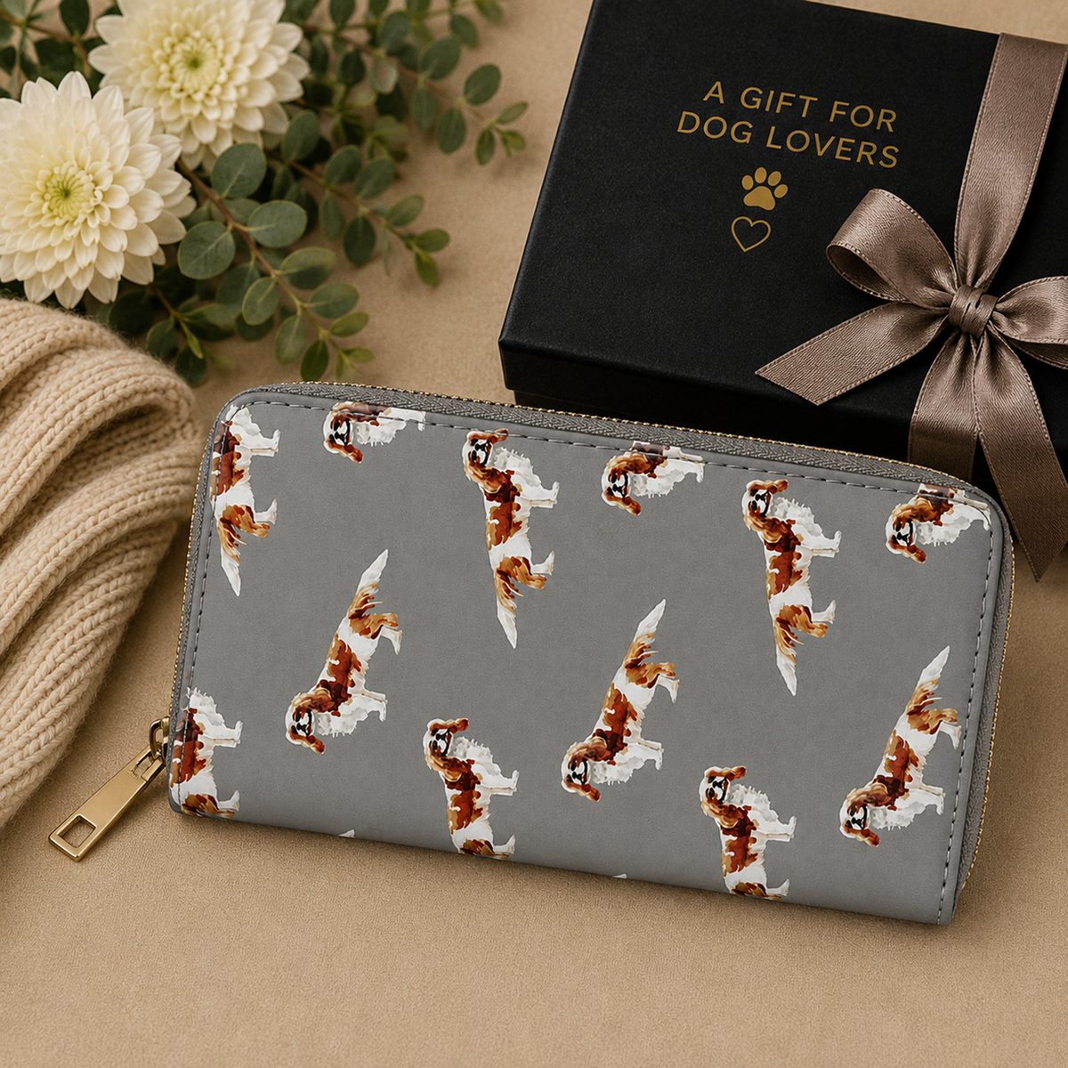 A grey zip-around wallet with a repeating pattern of small brown-and-white King Charles Cavalier Spaniel dogs, styled on a beige surface with a black gift box, flowers, and a knit blanket. The gift box has gold text reading 'A GIFT FOR DOG LOVERS'.