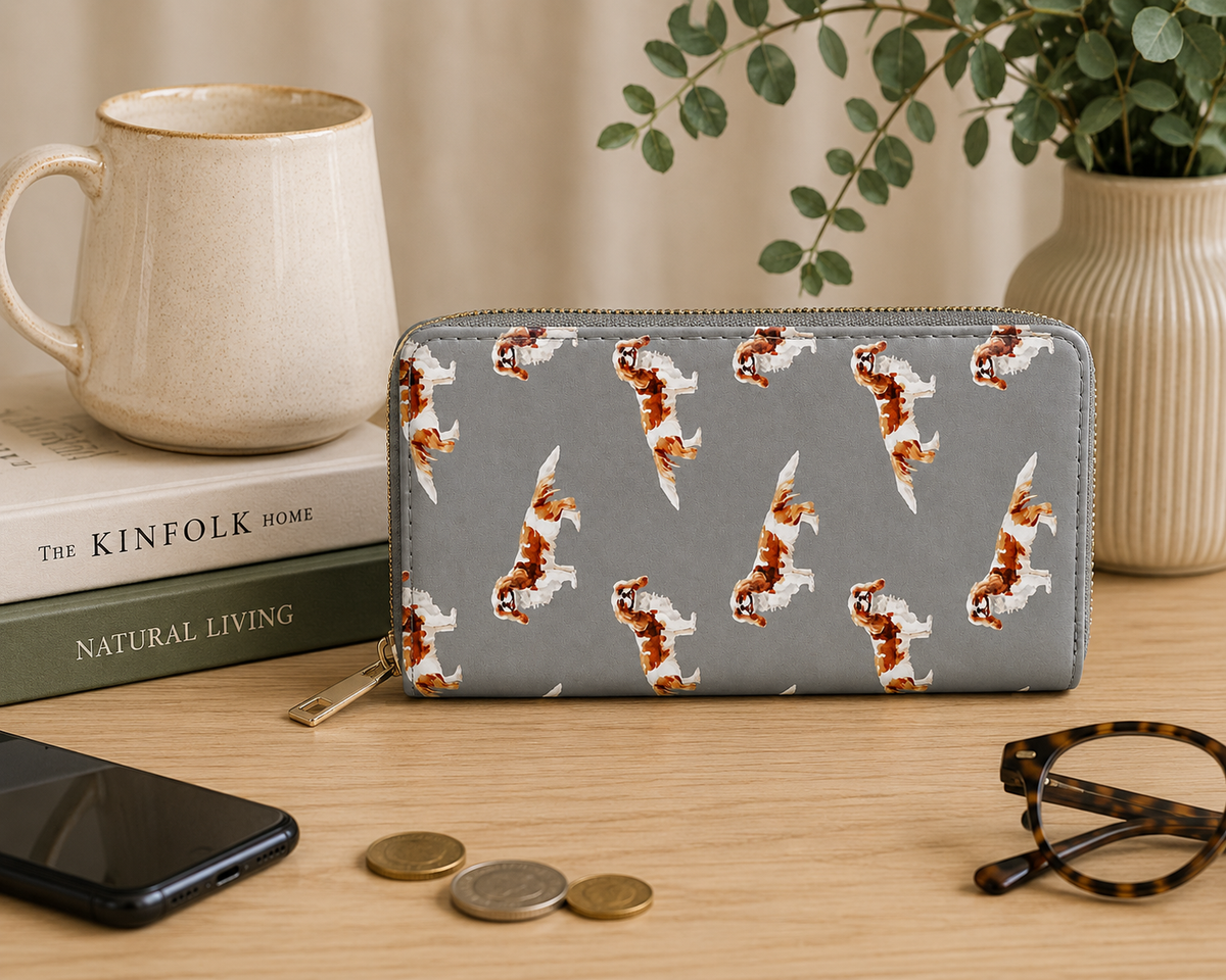 A grey zip-around wallet with a repeating pattern of small brown-and-white King Charles Cavalier Spaniel dog illustrations, styled on a wooden tabletop with a mug, books, phone, coins, and eyeglasses nearby.