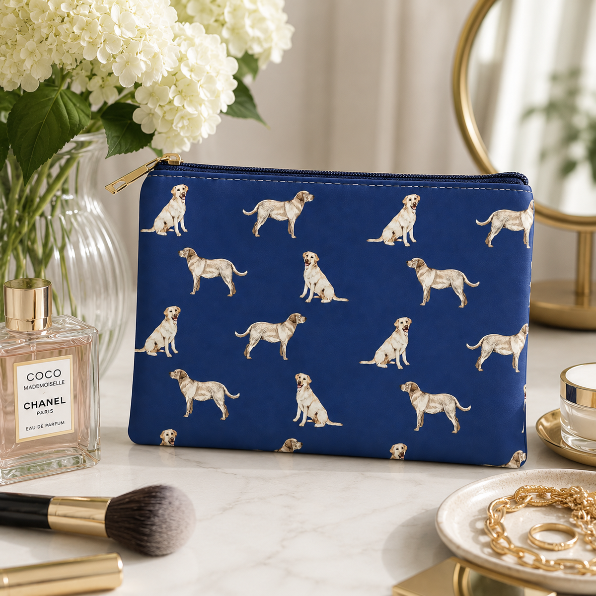 A navy blue zipped cosmetic bag with a repeating pattern of golden Labrador Retriever dogs, styled on a marble surface next to a Chanel perfume bottle, flowers, and gold accessories.